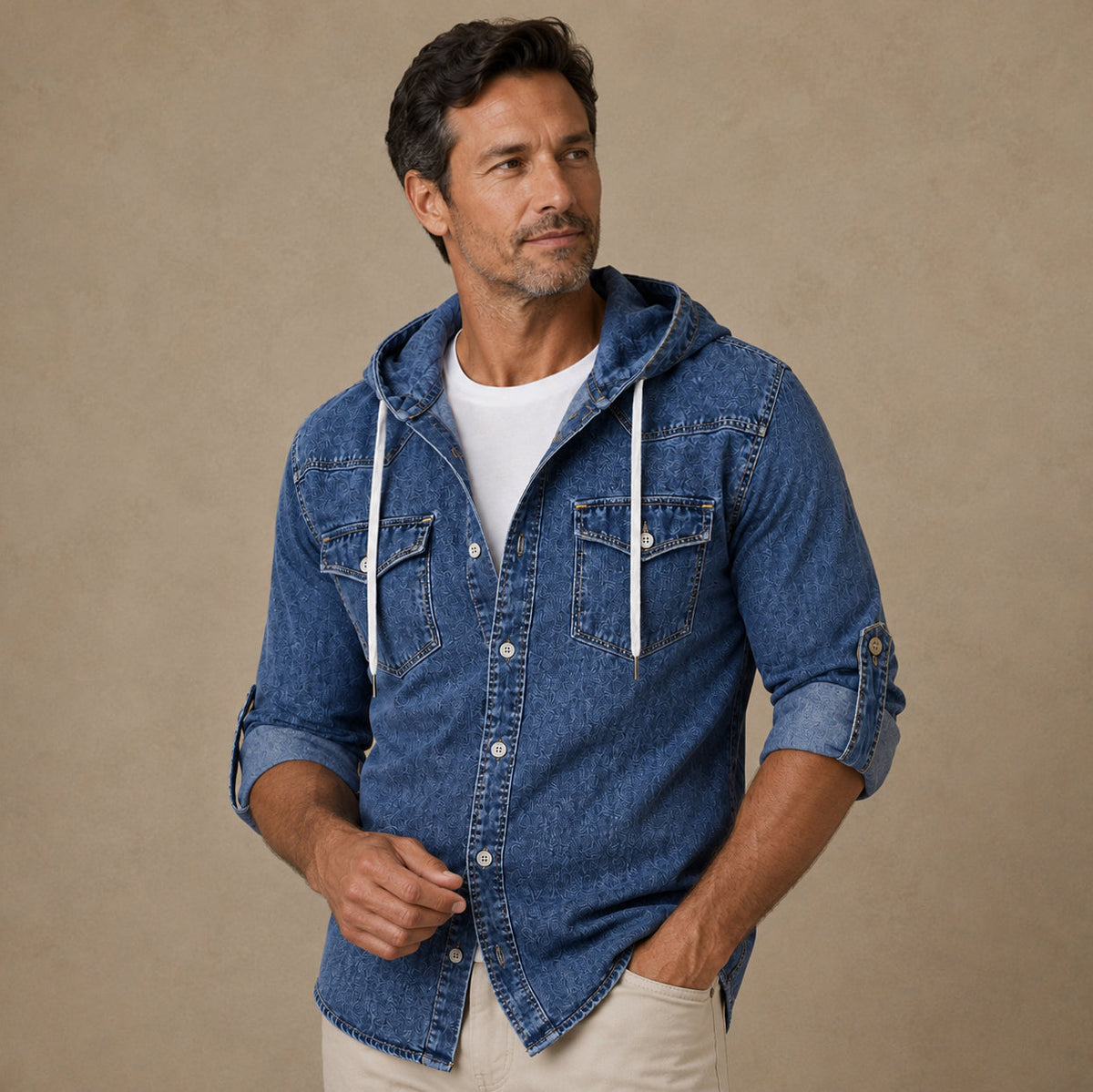 A man wearing a blue denim hooded button-up shirt over a white t-shirt and beige pants, standing against a textured beige studio background.