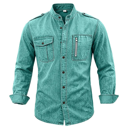 A men's long-sleeve button-up shirt in a washed teal color, featuring a mandarin collar, two chest flap pockets, and a vertical zippered pocket on the left chest, displayed against a textured gray background.