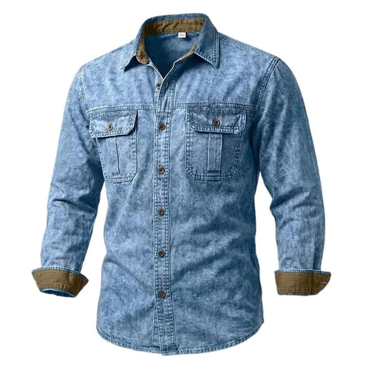 A men's long-sleeve button-up shirt in a washed blue denim-like fabric, featuring two chest flap pockets and brown button accents, with a contrasting brown inner collar and rolled cuffs, shown on a plain white background.