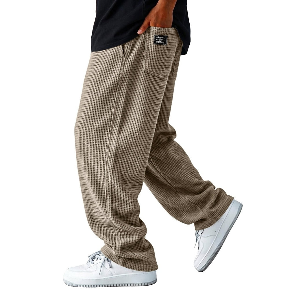 A person wearing loose, wide-leg brown and beige houndstooth pants with a black shirt and white sneakers, shown from the waist down on a plain white background.