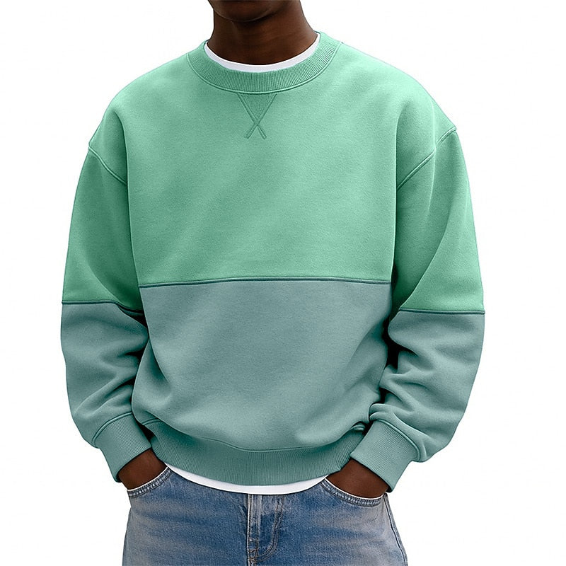 A person wearing a color-block crewneck sweatshirt with a mint green upper section and sleeves, and a muted sage green lower section, paired with blue jeans against a plain white background.
